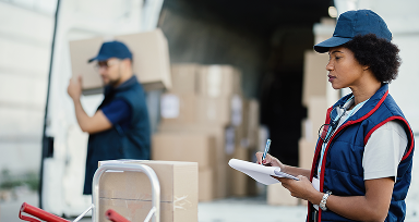 Vista de una mujer revisando entregas en cajas. Atrás hay un hombre llevando una caja y al fondo hay más cajas.