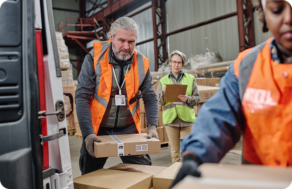 Hombre cargando una caja. Al frente se ve una mujer cargando otra caja. Al fondo se ve otra mujer y varias cajas.